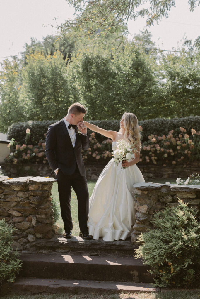 A groom in a black tuxedo gently kisses his bride’s hand as she smiles at him in a sunlit garden, surrounded by stone walls and blooming greenery during their wedding portraits.