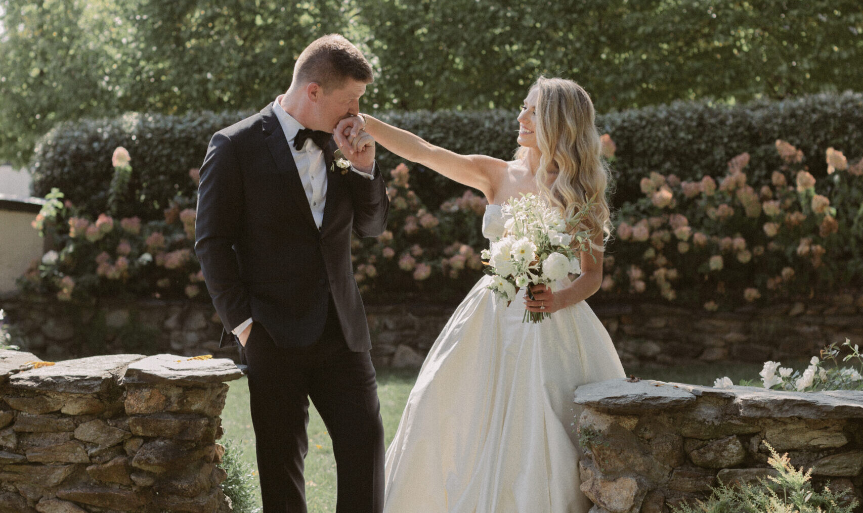 A groom in a black tuxedo gently kisses his bride’s hand as she smiles at him in a sunlit garden, surrounded by stone walls and blooming greenery during their wedding portraits.
