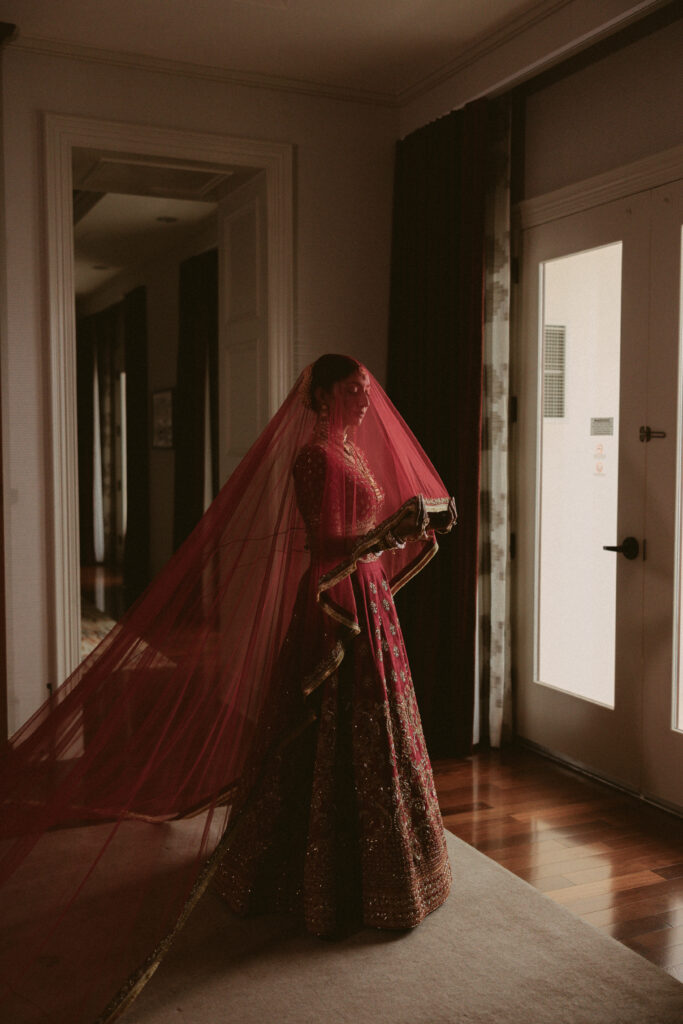 Bride wearing a red and gold South Asian bridal lehenga stands near a window with her long veil flowing behind her at the Gaylord National Resort.
