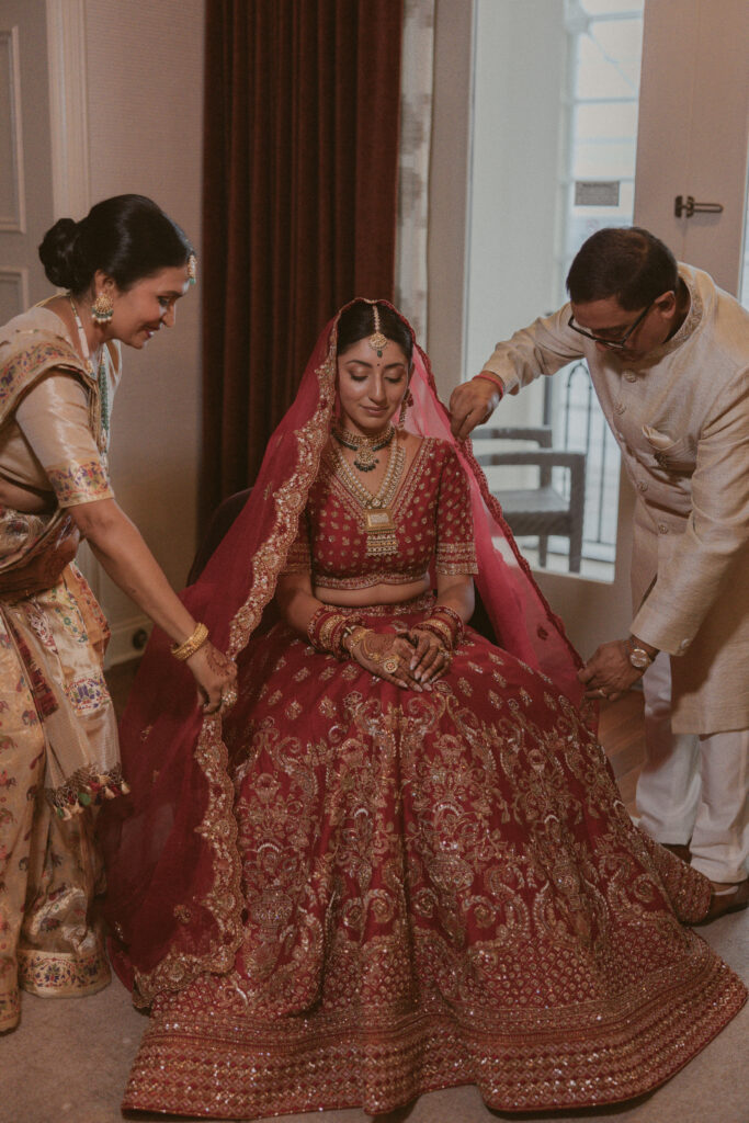South Asian bride sits in her red lehenga while her parents lovingly fix her veil before the ceremony at the Gaylord National Resort.